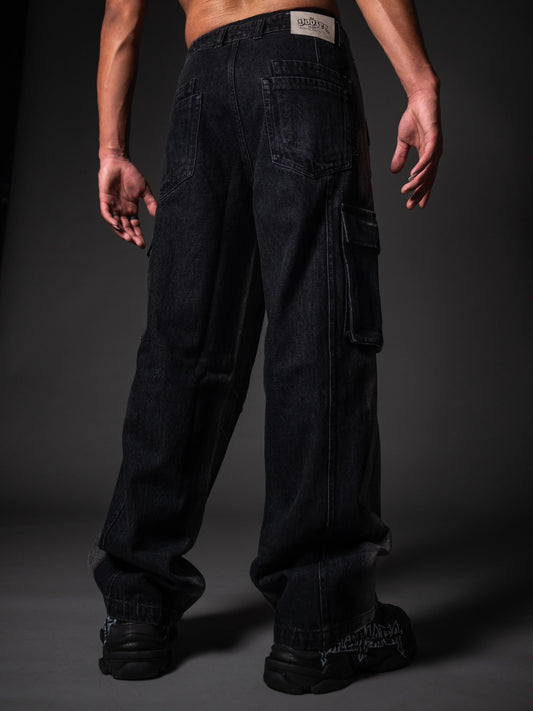 A back view of a model wearing loose-fitting, black denim cargo pants. The jeans feature standard back pockets, a side cargo pocket on the right leg, and a white branded patch on the right waistband. The look is completed with chunky black sneakers featuring white graphic details, set against a dark grey studio background.