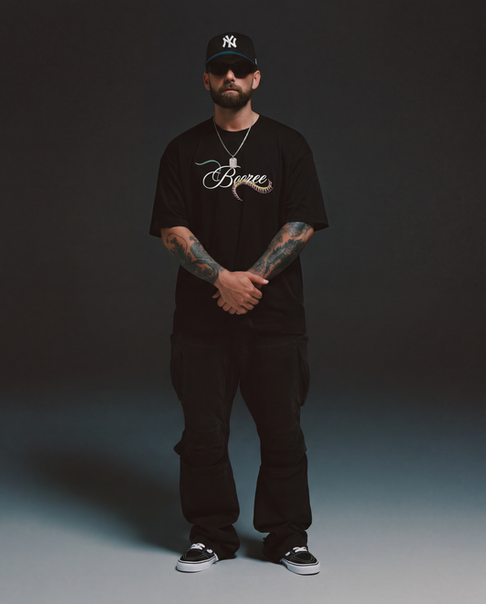 Man wearing a black t-shirt with a logo, black pants, and a black cap on a dark background