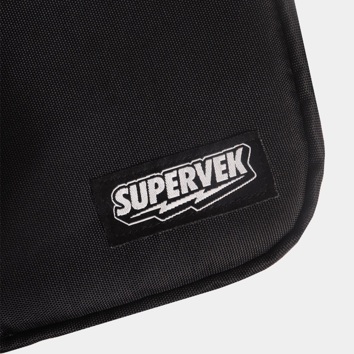 Detailed close-up of stitched Supervek logo patch on black "Sling" bag with textured fabric and Classic Black #color_Classic Black