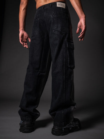A back view of a model wearing loose-fitting, black denim cargo pants. The jeans feature standard back pockets, a side cargo pocket on the right leg, and a white branded patch on the right waistband. The look is completed with chunky black sneakers featuring white graphic details, set against a dark grey studio background.