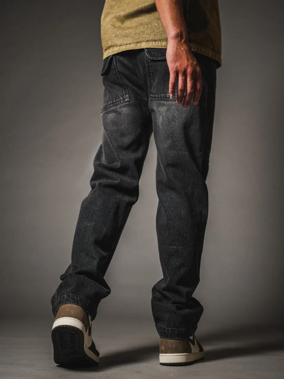 A back view of a model wearing relaxed-fit, faded black denim jeans. The jeans feature a vintage wash and distinctive back pockets with angled top stitching and flaps. The look is completed with an olive green shirt and low-top sneakers in black, cream, and brown, set against a dark grey studio background