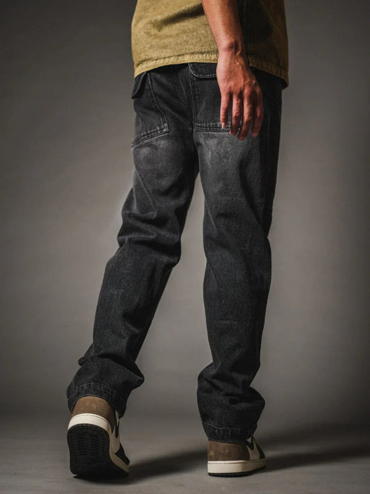 A back view of a model wearing relaxed-fit, faded black denim jeans. The jeans feature a vintage wash and distinctive back pockets with angled top stitching and flaps. The look is completed with an olive green shirt and low-top sneakers in black, cream, and brown, set against a dark grey studio background
