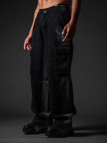 A model from the waist down wearing loose-fitting, faded black denim cargo pants with large side pocket flaps. The outfit is paired with chunky black sneakers featuring white graffiti-style lettering on the sides, set against a dark grey studio background.