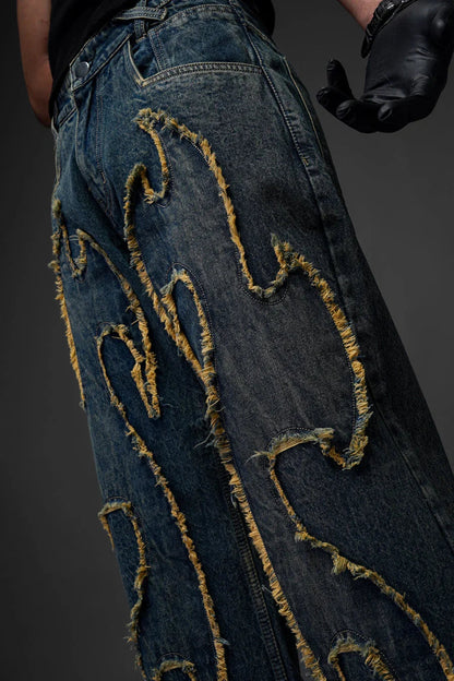 A close-up side view of a person wearing dark blue denim jeans. The focus is on the intricate, raised appliquÃ© design that mimics flames, featuring heavily frayed, yellowish-tan edges. A hand wearing a black leather glove is visible on the right side of the frame.