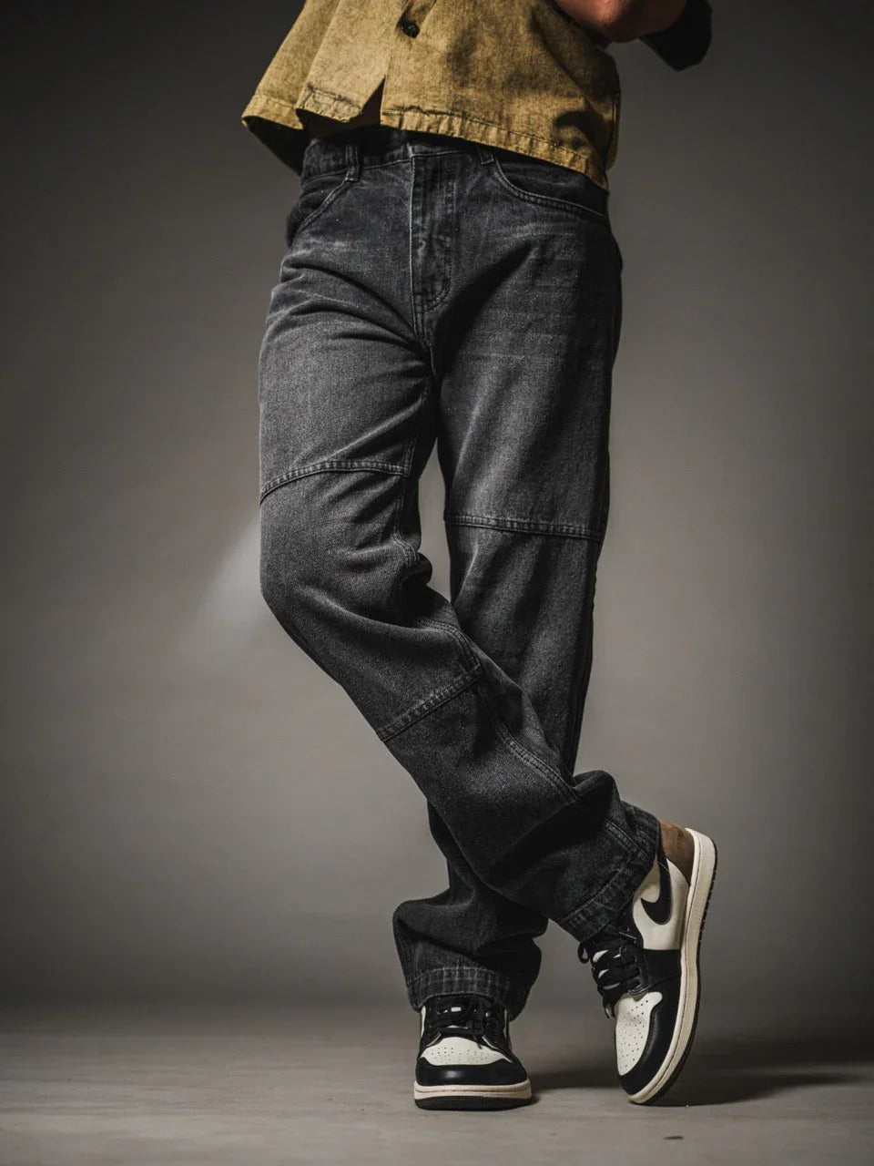 A person from the waist down wearing faded black denim jeans with horizontal seam panels across the knees. They are standing with their legs casually crossed at the ankles. The outfit includes a textured olive-green shirt and black and cream low-top sneakers, set against a dark grey studio background.