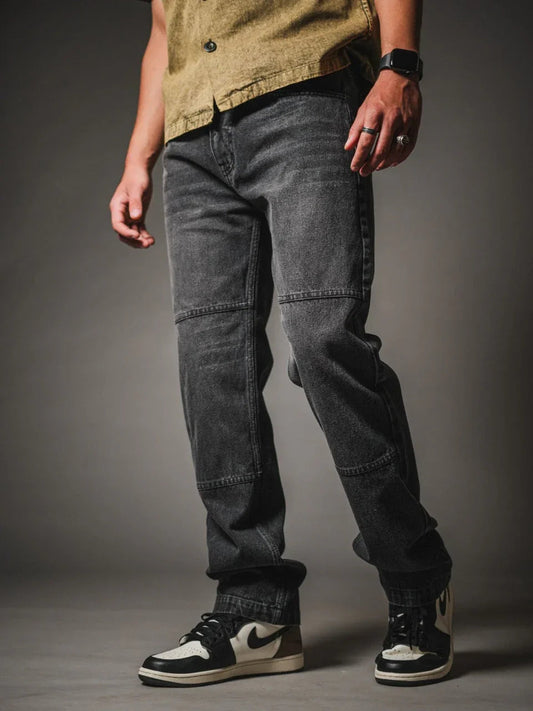 A model from the chest down wearing faded black, straight-leg denim jeans featuring horizontal seam panels across the knees. The jeans are styled with a textured olive-green button-up shirt and black and cream low-top sneakers, set against a dark grey studio background.