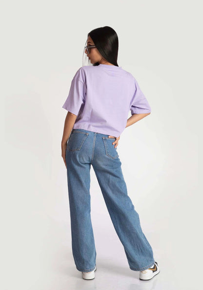 Back view of a woman wearing Stylera lavender oversized crop top with blue wide-leg jeans and sunglasses