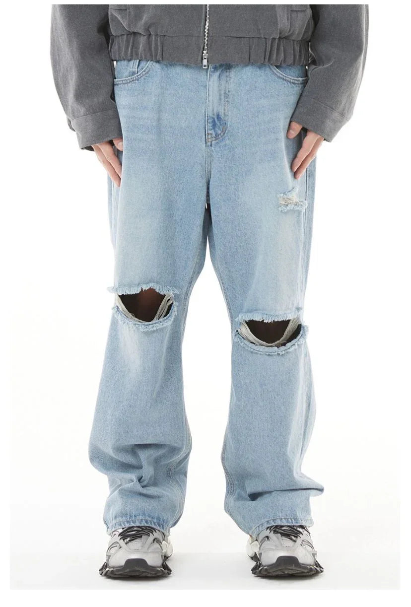 A direct, front-on photograph of a person against a plain white studio background, showcasing a streetwear outfit. The main focus is the pair of baggy, light-wash blue denim jeans, which feature large, clean-cut, horizontal rips across both knees and a smaller distressed patch on the upper left thigh. Above the jeans, a charcoal grey, cropped, zip-up bomber jacket with a wide elasticated hem is worn. On the feet are chunky, multi-layered silver and black technical sneakers with thick soles. The perspective 