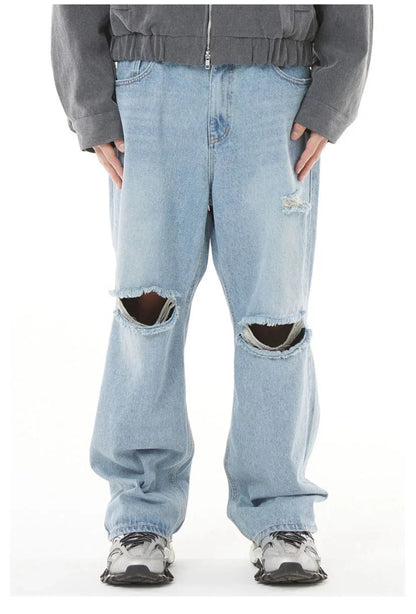 A direct, front-on photograph of a person against a plain white studio background, showcasing a streetwear outfit. The main focus is the pair of baggy, light-wash blue denim jeans, which feature large, clean-cut, horizontal rips across both knees and a smaller distressed patch on the upper left thigh. Above the jeans, a charcoal grey, cropped, zip-up bomber jacket with a wide elasticated hem is worn. On the feet are chunky, multi-layered silver and black technical sneakers with thick soles. The perspective 