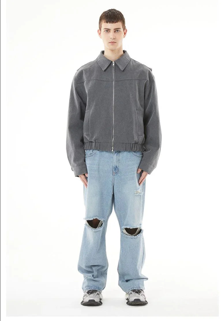 A full-length, front-facing product shot of a young male model with short hair, standing against a plain white background. He is wearing a charcoal grey, zippered canvas jacket with a collar and an elasticated waistband, paired with light-wash, wide-leg blue jeans. The jeans feature prominent, wide horizontal rips and distressed details at both knees, showing part of his legs. On his feet are chunky grey and white multi-layered sneakers. He stands straight with a neutral expression.