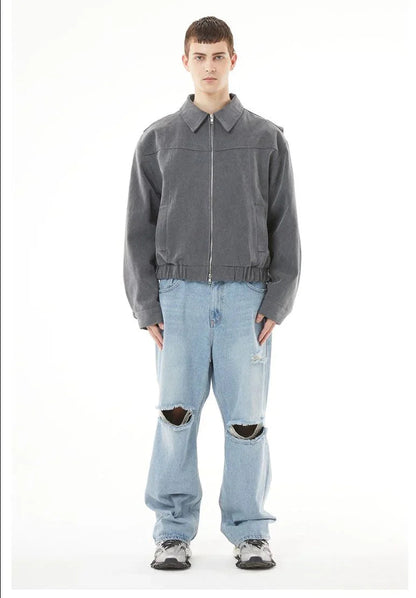 A full-length, front-facing product shot of a young male model with short hair, standing against a plain white background. He is wearing a charcoal grey, zippered canvas jacket with a collar and an elasticated waistband, paired with light-wash, wide-leg blue jeans. The jeans feature prominent, wide horizontal rips and distressed details at both knees, showing part of his legs. On his feet are chunky grey and white multi-layered sneakers. He stands straight with a neutral expression.