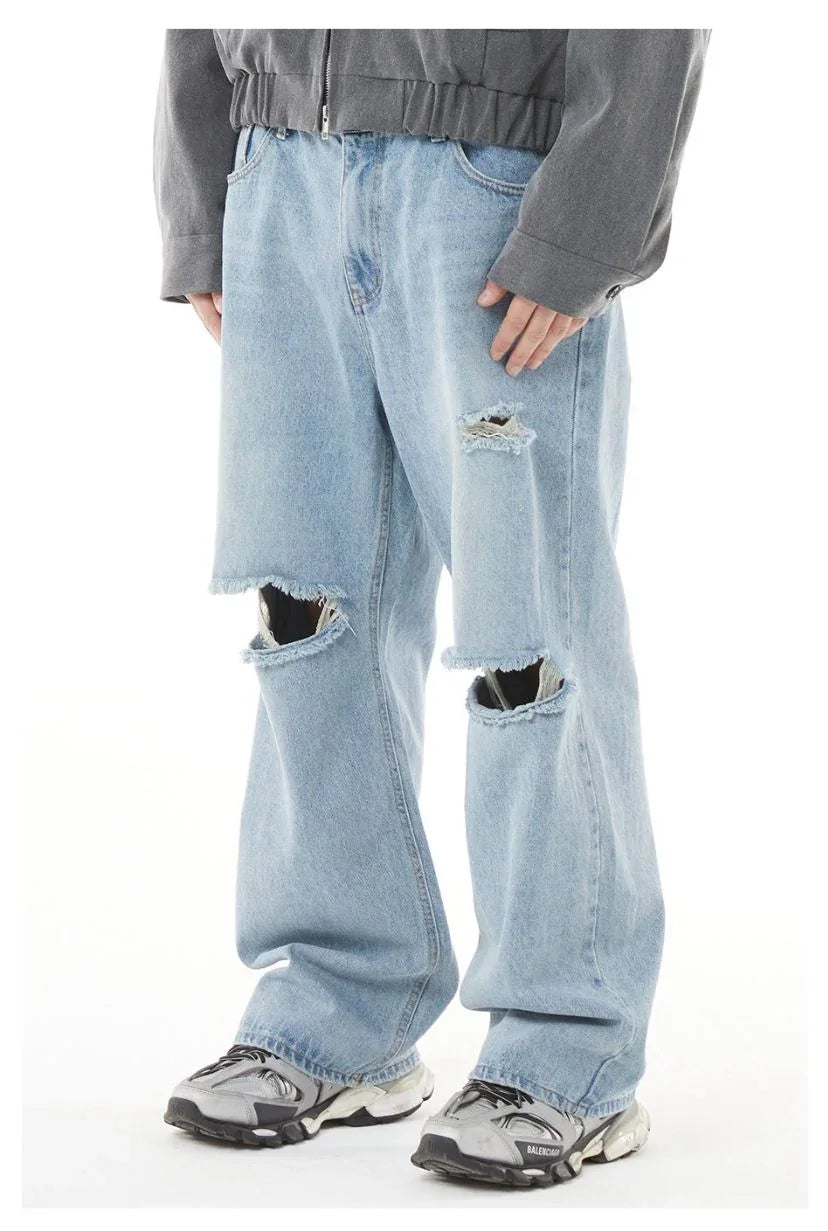 A model standing against a solid white background wearing light-wash blue baggy denim jeans. The jeans feature prominent distressed, large horizontal rips across both knees and a smaller distressed patch on the upper left thigh. They are styled with a grey cropped zip-up jacket and chunky grey and black sneakers.
