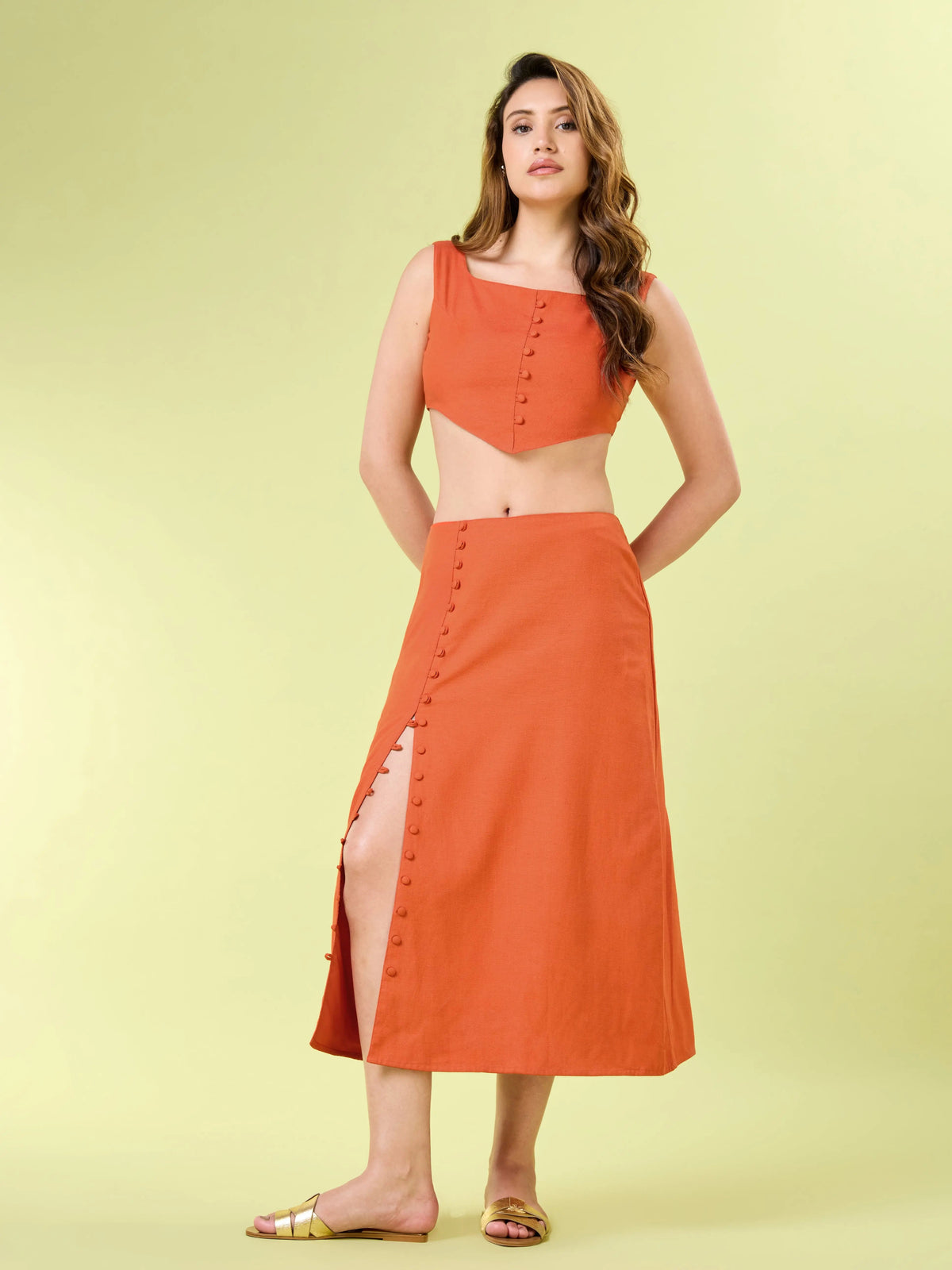 Full body shot of a woman modeling an orange two-piece co-ord set. She is wearing a square-neck crop top with a pointed hem and a matching midi skirt with button detailing that opens into a high side slit. She is styled with flat gold sandals.