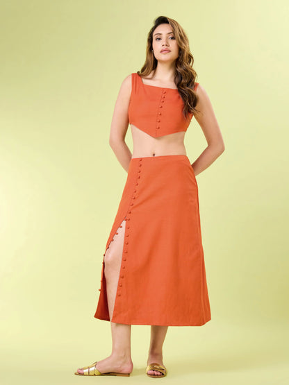 Full body shot of a woman modeling an orange two-piece co-ord set. She is wearing a square-neck crop top with a pointed hem and a matching midi skirt with button detailing that opens into a high side slit. She is styled with flat gold sandals.